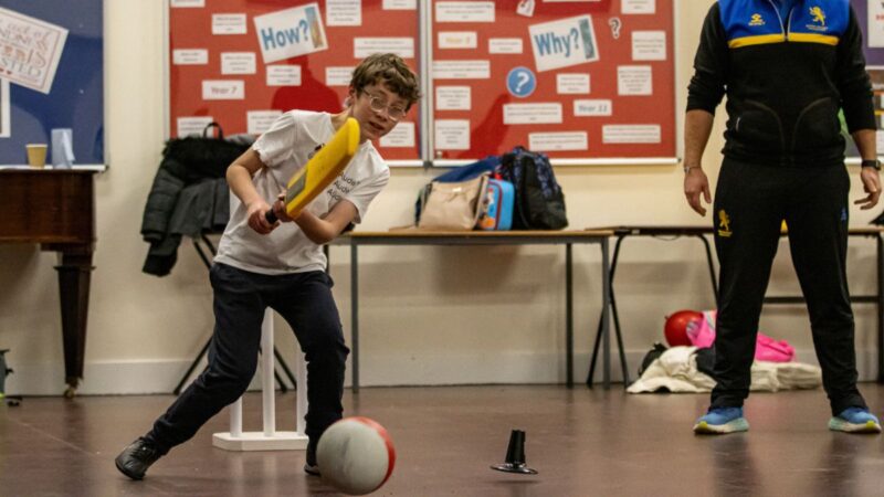 Boy in the classroom playing, (supervised) with a cricket bat and a football, having fun.