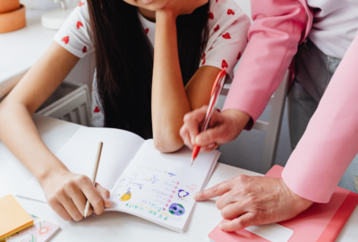 An adult points to a child’s notebook filled with drawings while the child sits at a table.