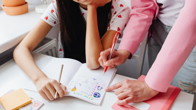 An adult points to a child’s notebook filled with drawings while the child sits at a table.