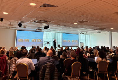 Large group of people seated in a conference room listening to a speaker on stage with two screens.