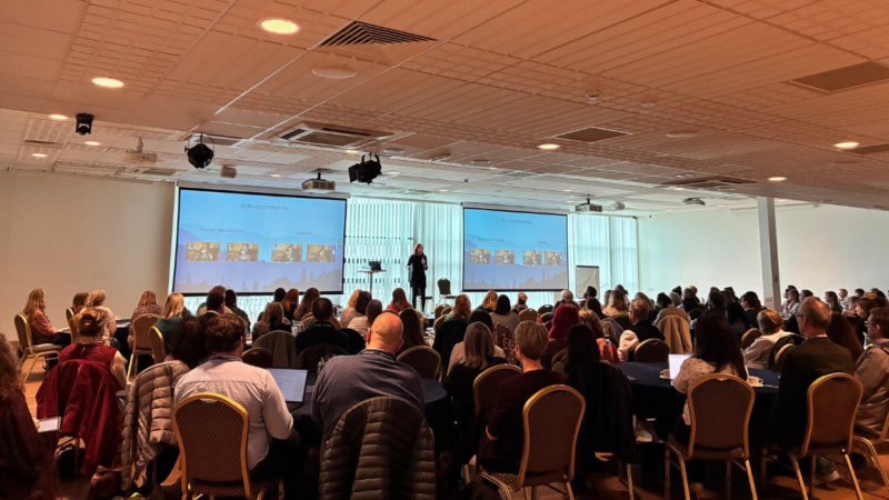 Large group of people seated in a conference room listening to a speaker on stage with two screens.