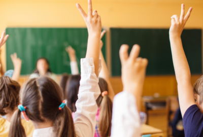 Children sitting in a classroom with their hands raised, facing a teacher at the front of the room.