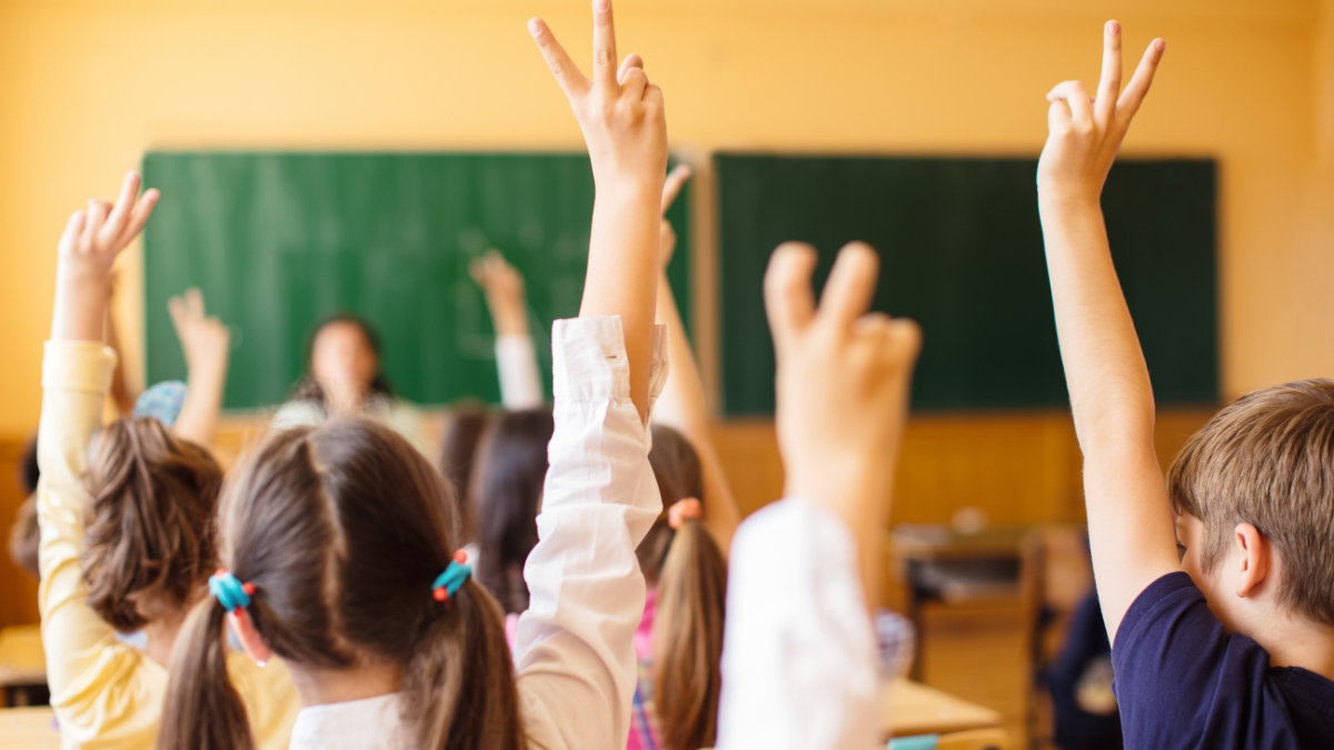 Children sitting in a classroom with their hands raised, facing a teacher at the front of the room.