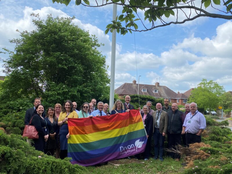 Group of staff standing outdoors around a flagpole, holding a rainbow Pride flag with the Devon County Council logo, with trees, houses and a blue sky in the background.