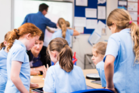 A group of school children working with a teaching assistant around a table. In the background, two other pupils are working with a teacher on a whiteboard.