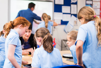 A group of school children working with a teaching assistant around a table. In the background, two other pupils are working with a teacher on a whiteboard.