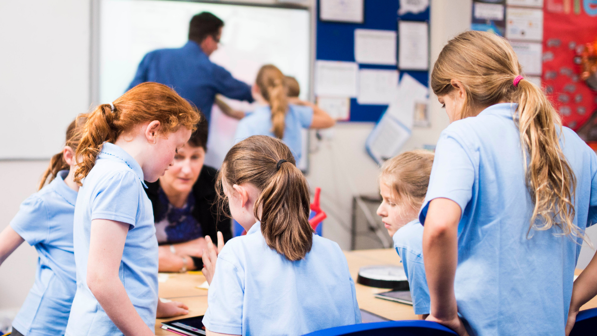 A group of school children working with a teaching assistant around a table. In the background, two other pupils are working with a teacher on a whiteboard.
