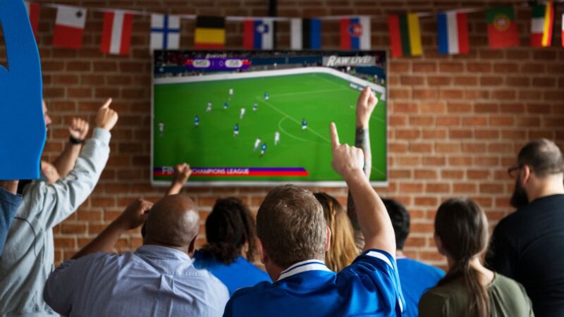 Adults watching a big screen with the football on. Men waving their arms