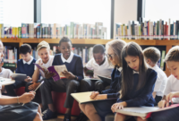 A group of junior shcoolchildren sitting in a library using a range of books and devices to read