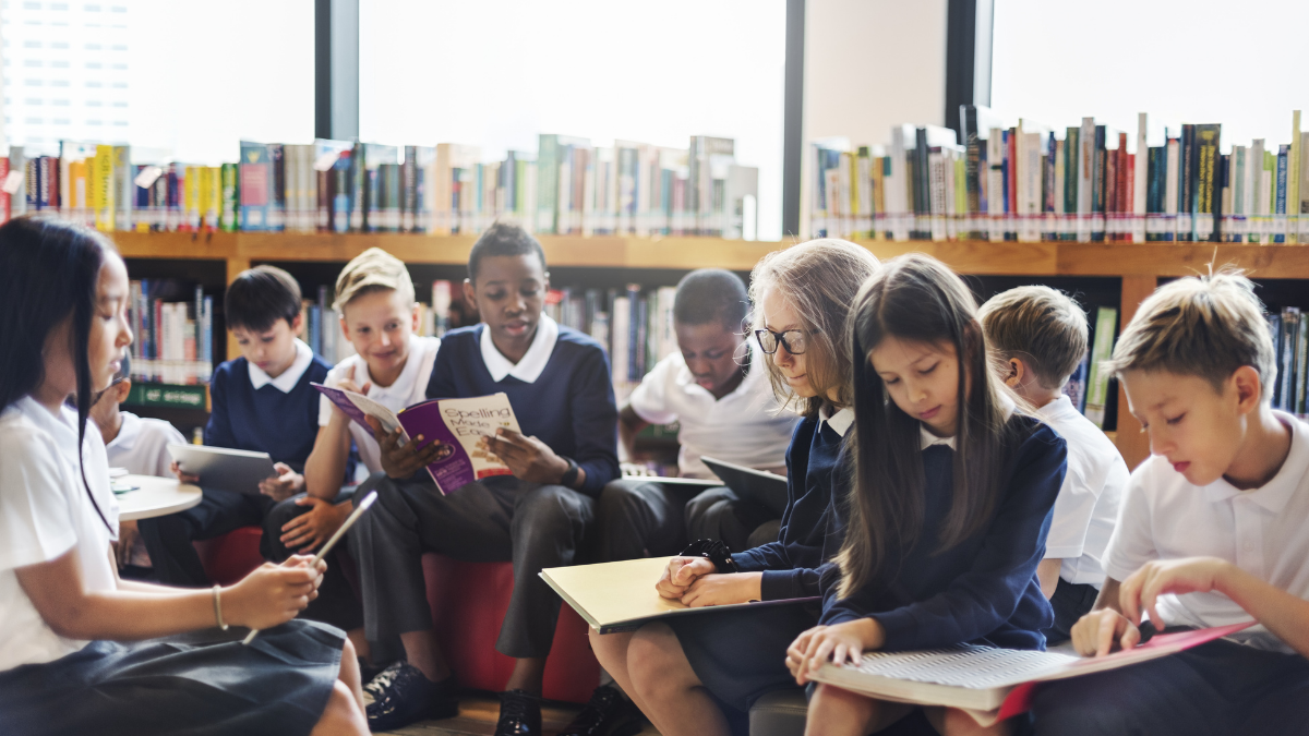 A group of junior shcoolchildren sitting in a library using a range of books and devices to read