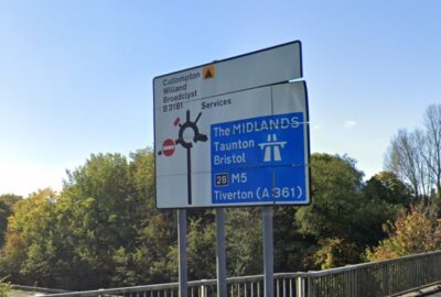 Road sign near Cullompton indicating routes to the M5, Tiverton, Taunton, Bristol, Willand and Broadclyst, with trees and blue sky behind.