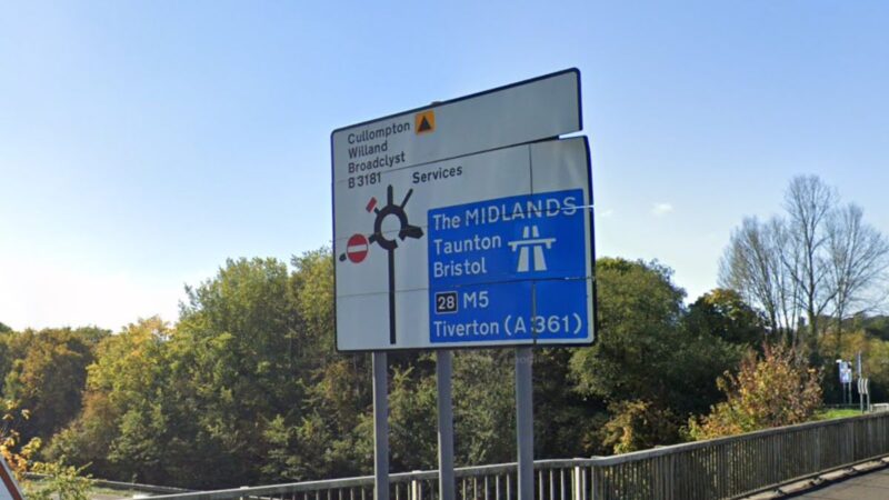 Road sign near Cullompton indicating routes to the M5, Tiverton, Taunton, Bristol, Willand and Broadclyst, with trees and blue sky behind.