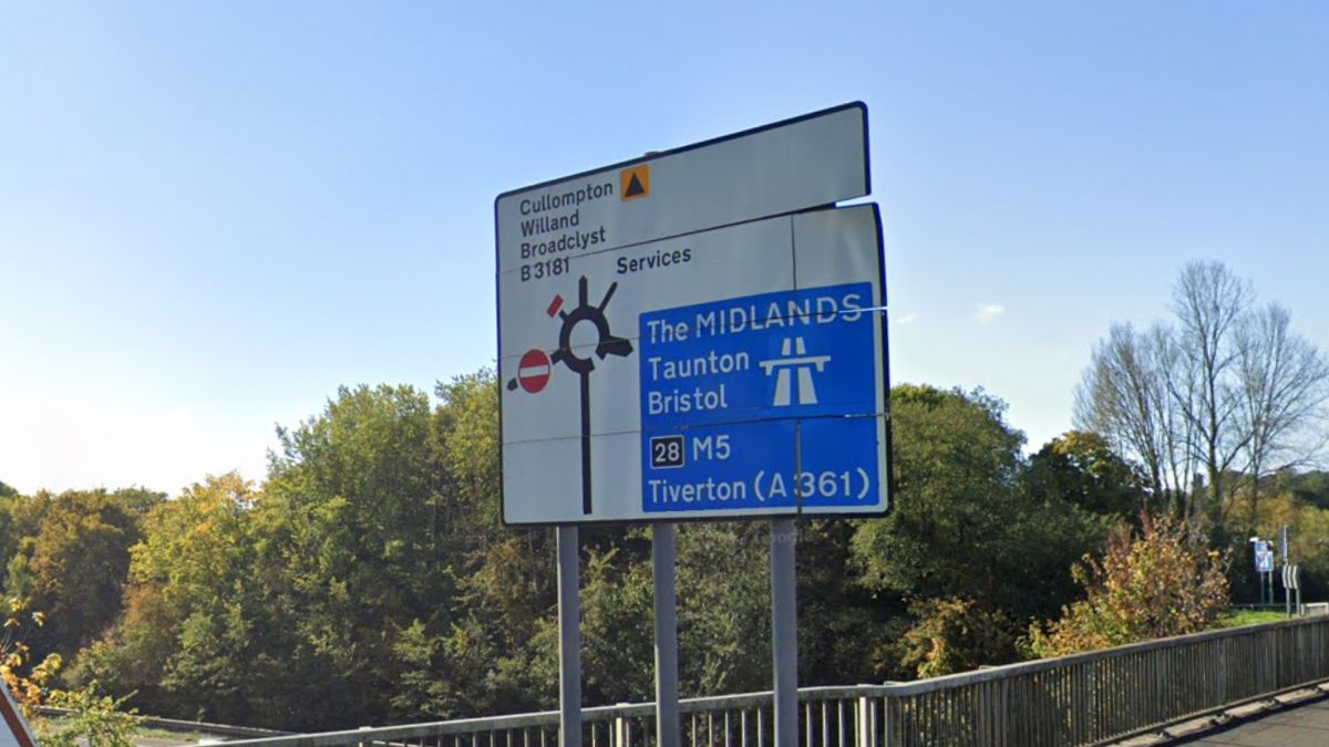 Road sign near Cullompton indicating routes to the M5, Tiverton, Taunton, Bristol, Willand and Broadclyst, with trees and blue sky behind.
