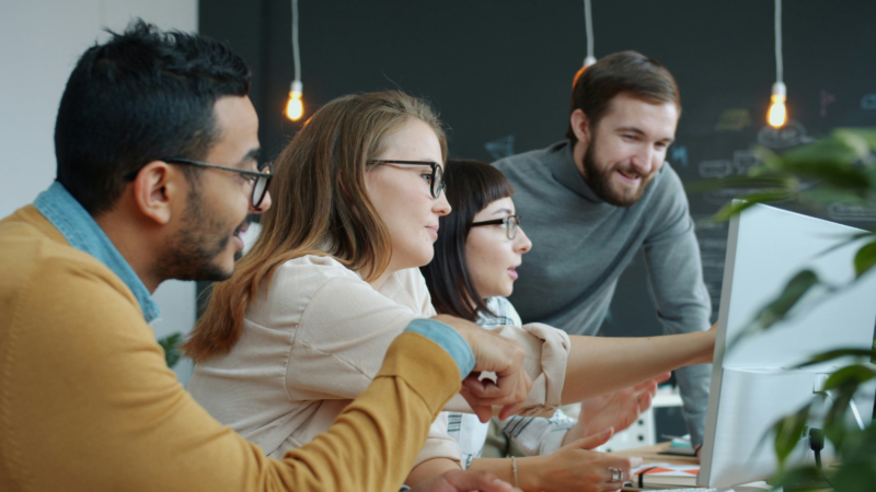 Group of young professionals looking at a computer screen together in a workplace environment.