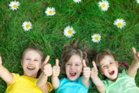 Three young children lay on their backs on the grass smiling at the camera and holding their thumbs up.