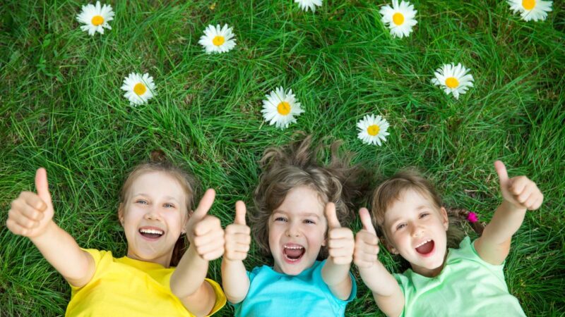 Three young children lay on their backs on the grass smiling at the camera and holding their thumbs up.