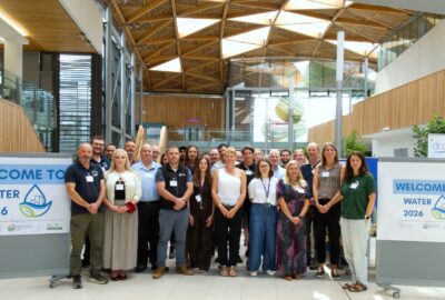 A large group of WATER 2026 representatives pose together between two conference welcome signs in the timber-roofed Forum at the University of Exeter.