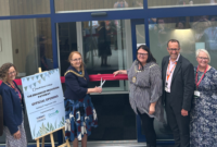 A group of people smiling as a woman wearing mayoral chains cuts the ribbon over a modern building