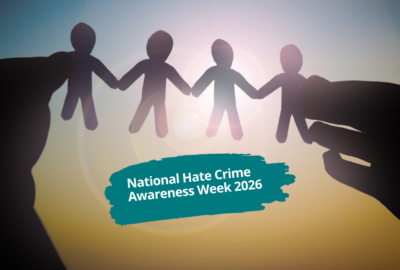 Silhouetted hands hold a chain of four paper figures against a sunlit background, with the text: “National Hate Crime Awareness Week 2026”.