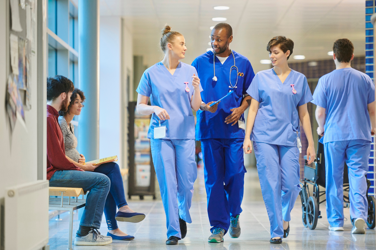 Three healthcare workers in blue scrubs walk and talk together along a hospital corridor, with other people in the background.