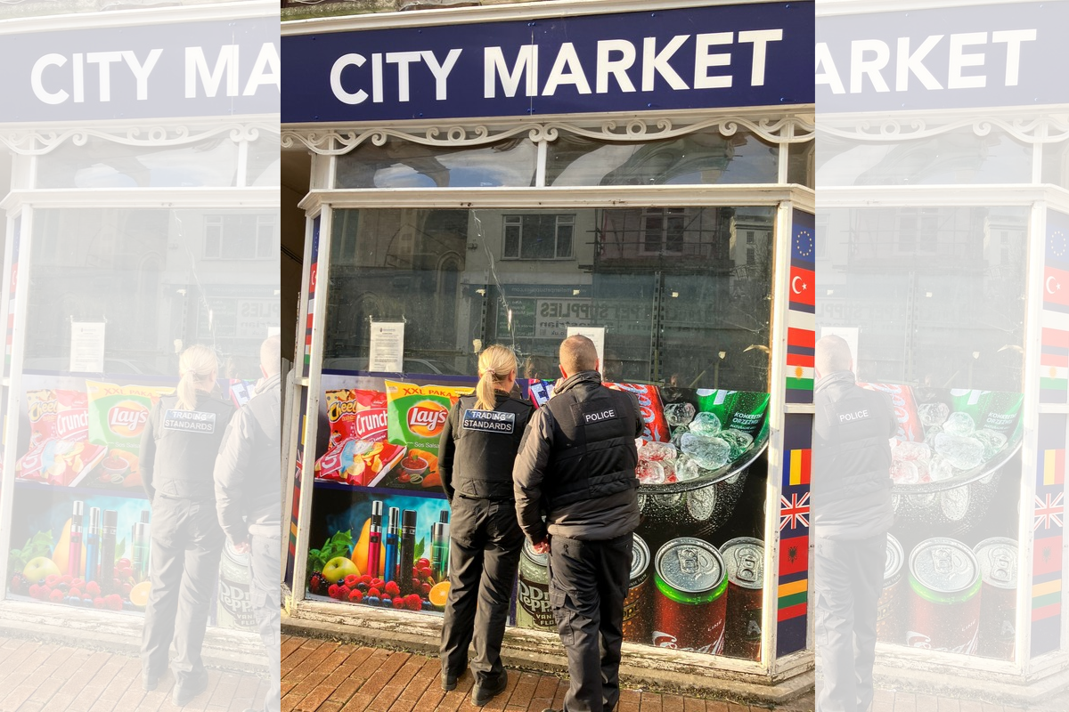 Two Trading Standards officers and a police officer stand outside a City Market shop, looking through its front window. Colourful advertisements for snacks, drinks and vaping products cover the lower part of the shopfront.
