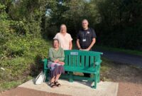 Three representatives from Devon County Council and Devon and Somerset Fire and Rescue Service beside a green mental health support bench on a woodland path.
