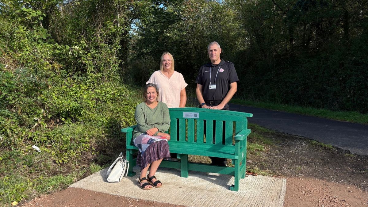 Three representatives from Devon County Council and Devon and Somerset Fire and Rescue Service beside a green mental health support bench on a woodland path.