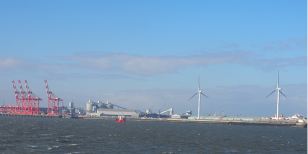 Wind turbines at Liverpool Docks. There is water in the foreground and the turbines can be seen alongside other tall structures.