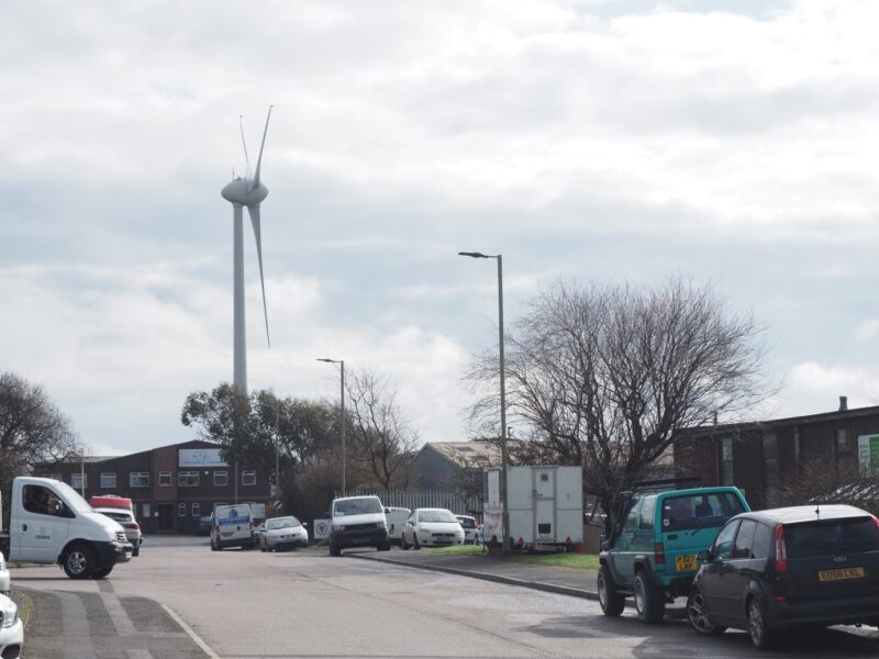 The wind turbine at Mullacott Cross industrial estate. The image contains a road with parked vehicles in the foreground and some warehouse style buildings.