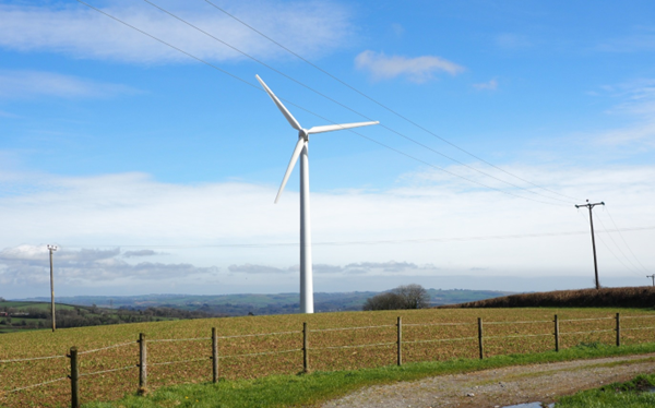 The South Brent Community Energy wind turbine. The turbine is in a field with countryside in the foreground and background and a blue sky.