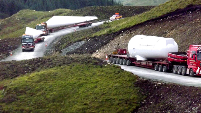 Three large lorries transporting the parts of wind turbines up a narrow road cut into the hillside.