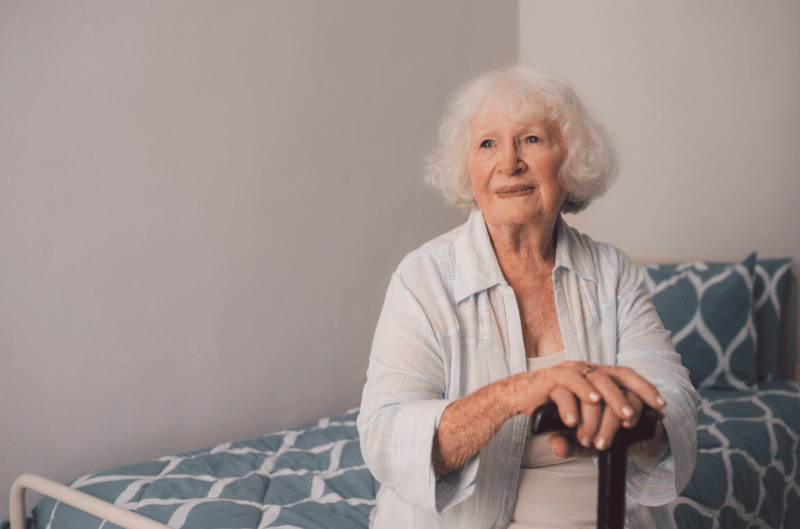 Older woman sitting on the edge of her bed with her hand on a walking stick: Replacement care