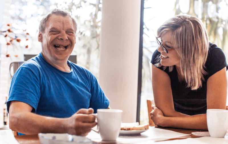 Man (who has a learning disability) laughing with his female supported living manager, sharing a smile over a mug of tea and breakfast.