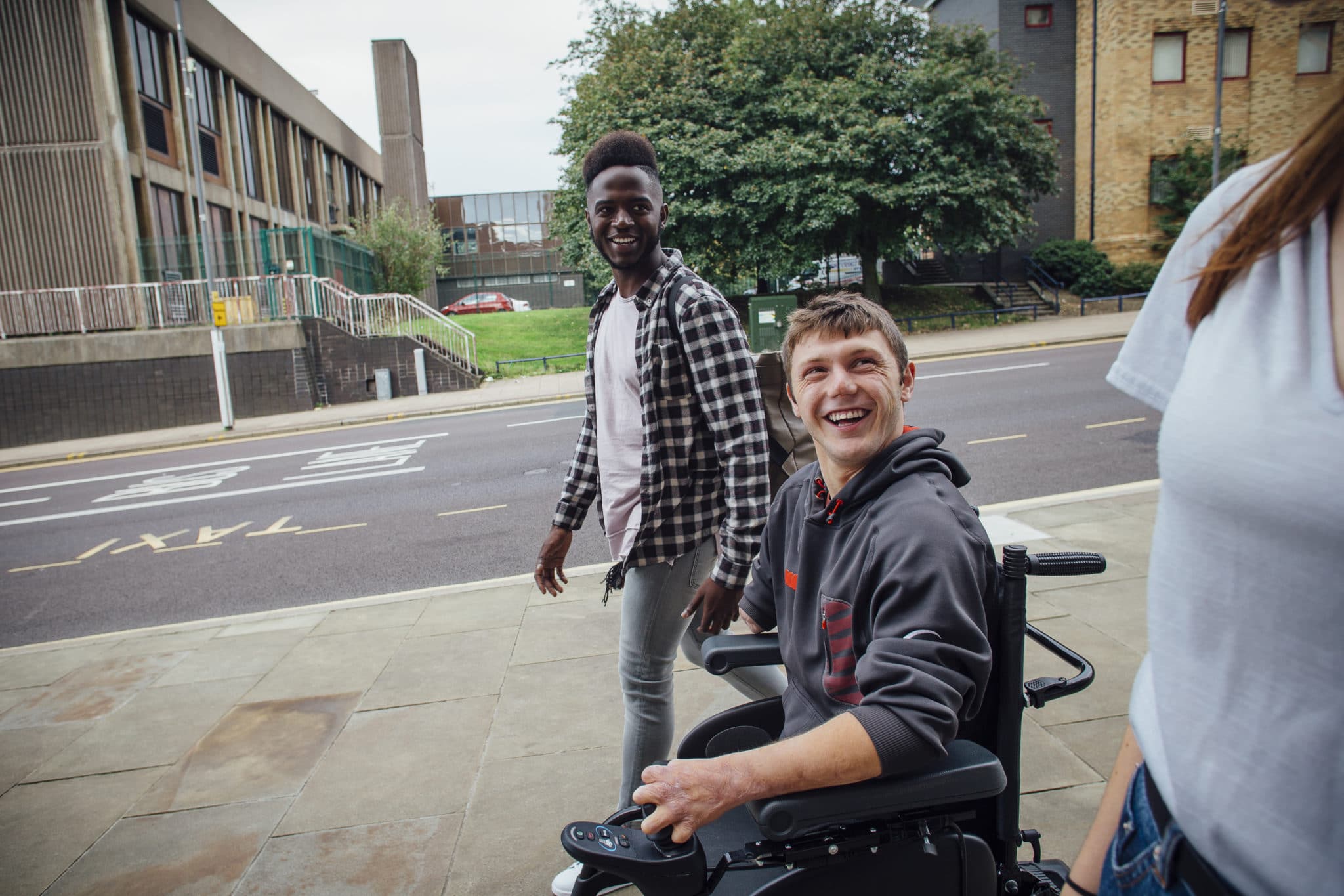 Three young adults on a pavement, one in a wheelchair, looking happy