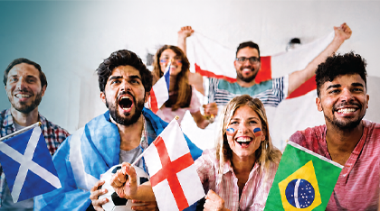 A diverse group of six soccer fans cheer enthusiastically while watching a match, waving national flags from Scotland, England, Brazil, and another country. The fans smile, shout, and celebrate together in a festive indoor setting, with face paint and team colours adding to the excitement.