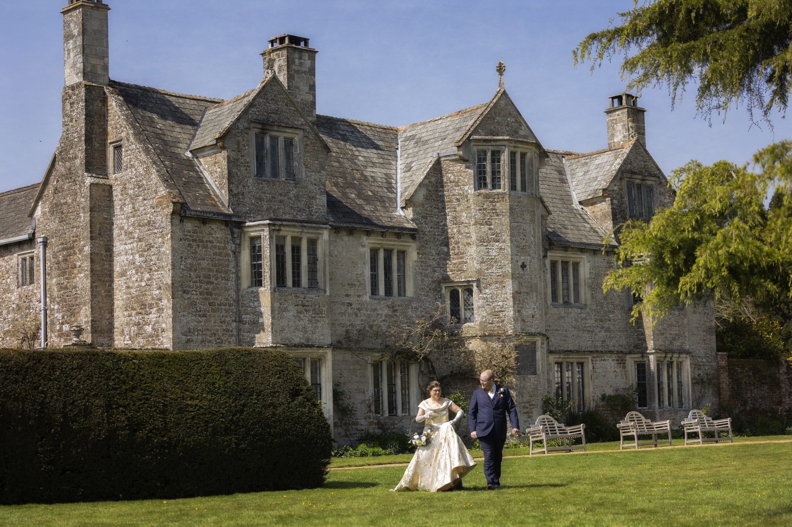 A bride and groom walking across a lawn with a large stone building behind them