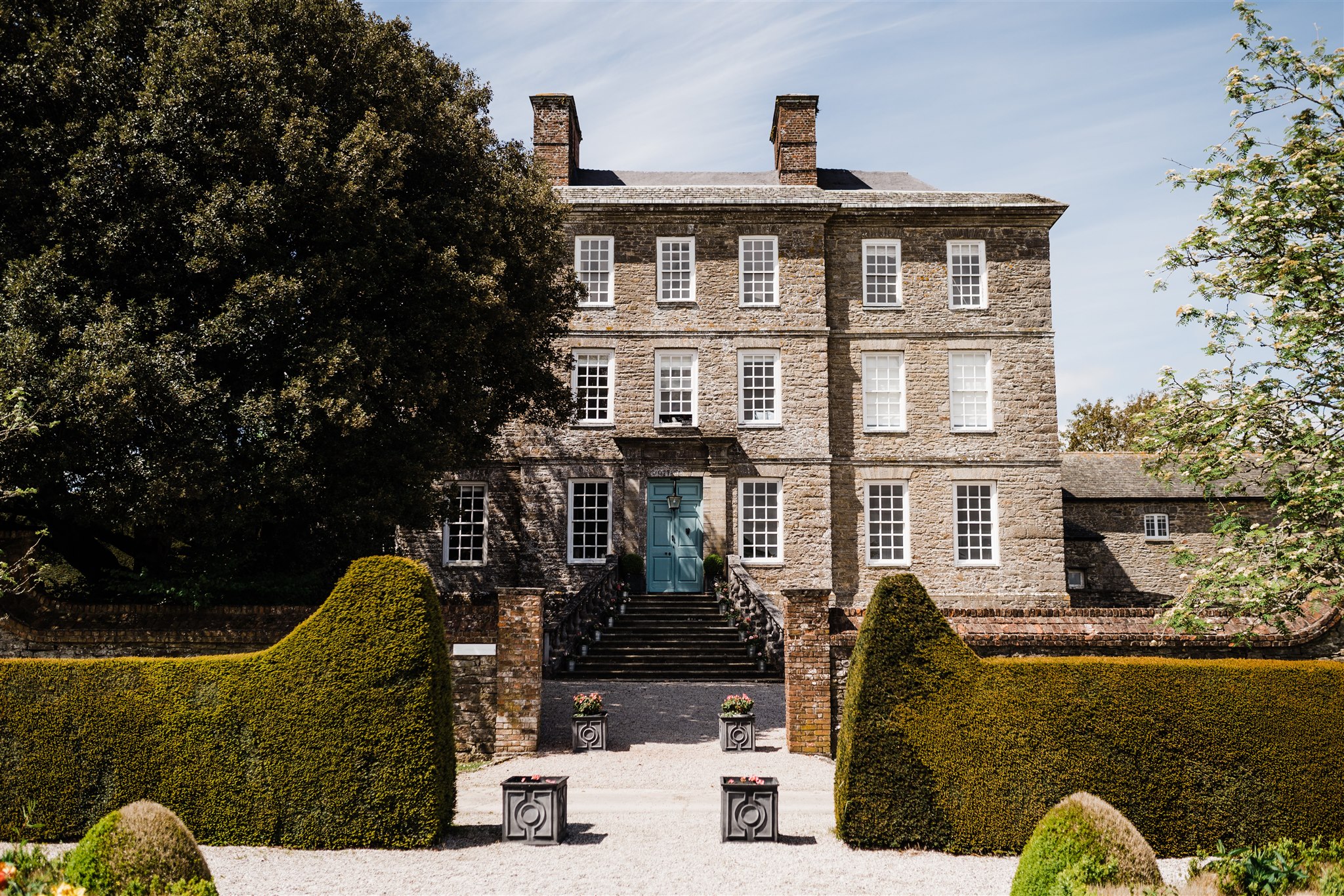 A large Georgian style building, with a stone staircase leading to the front door, with hedges and gardens framing the building