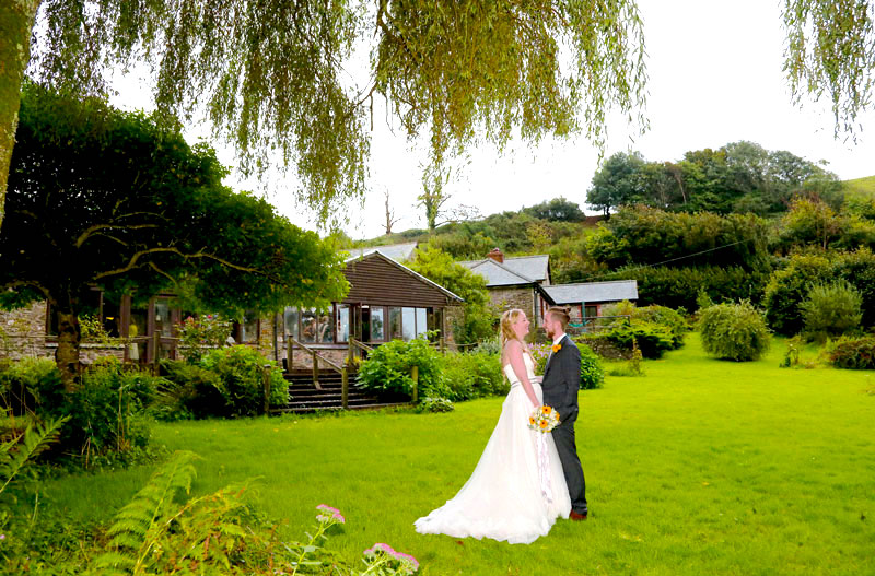 A bride and groom standing in gardens with a low building behind them.