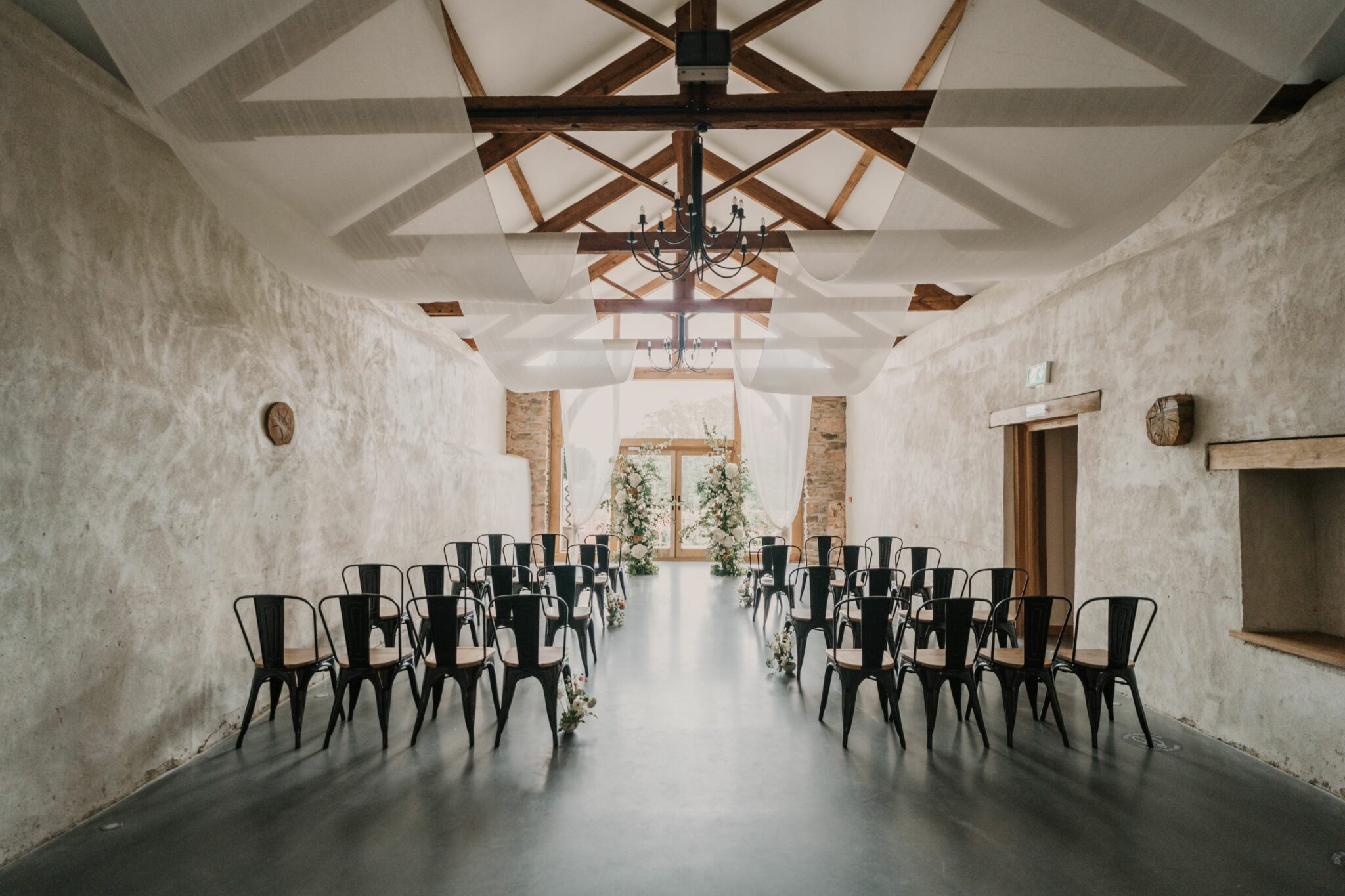 The inside of the barn, with a wooden beamed ceiling and pale walls. Rows of chairs are facing the front, which is decorated with flowers.