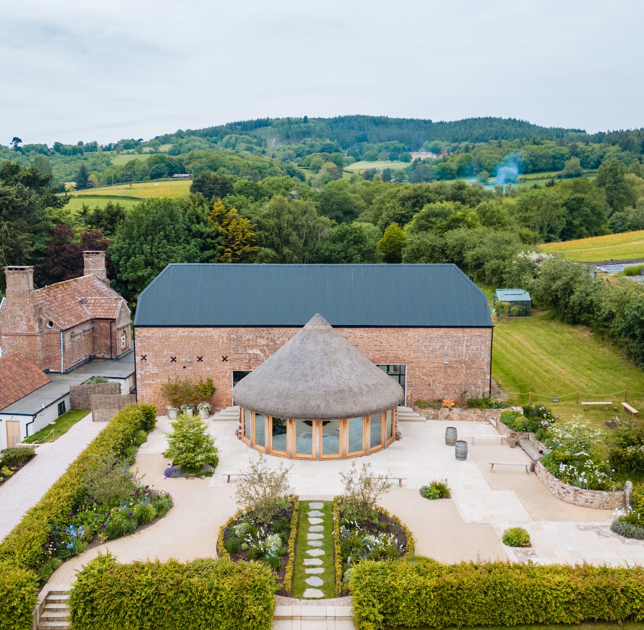 Aerial photo of a large brick building, with a circular building at the back, which has a coned, thatched roof. The buildings are set in a rural location.