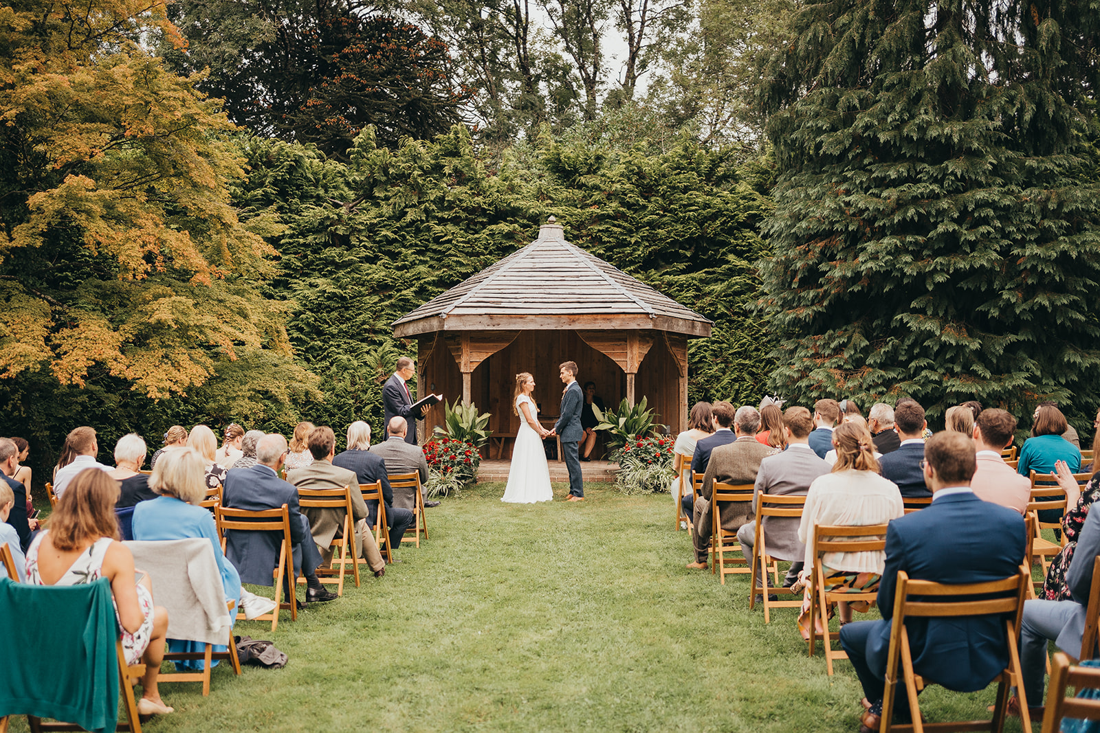 A bridge and groom standing in front of a wooden pergola, with rows of guests sitting facing them.