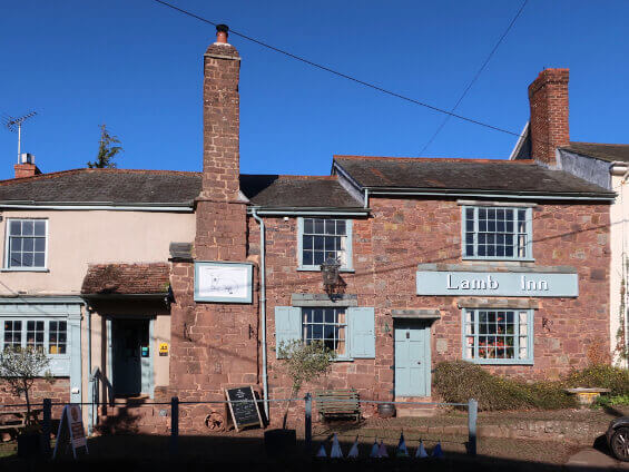A sandstone building, with tall chimneys, pale green wooden door, window frames, shutters and a sign saying The Lamb Inn.