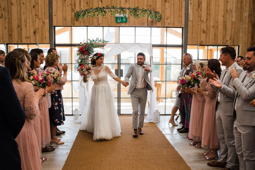 A bride and groom holding hands, with their weeding guests facing towards them and clapping