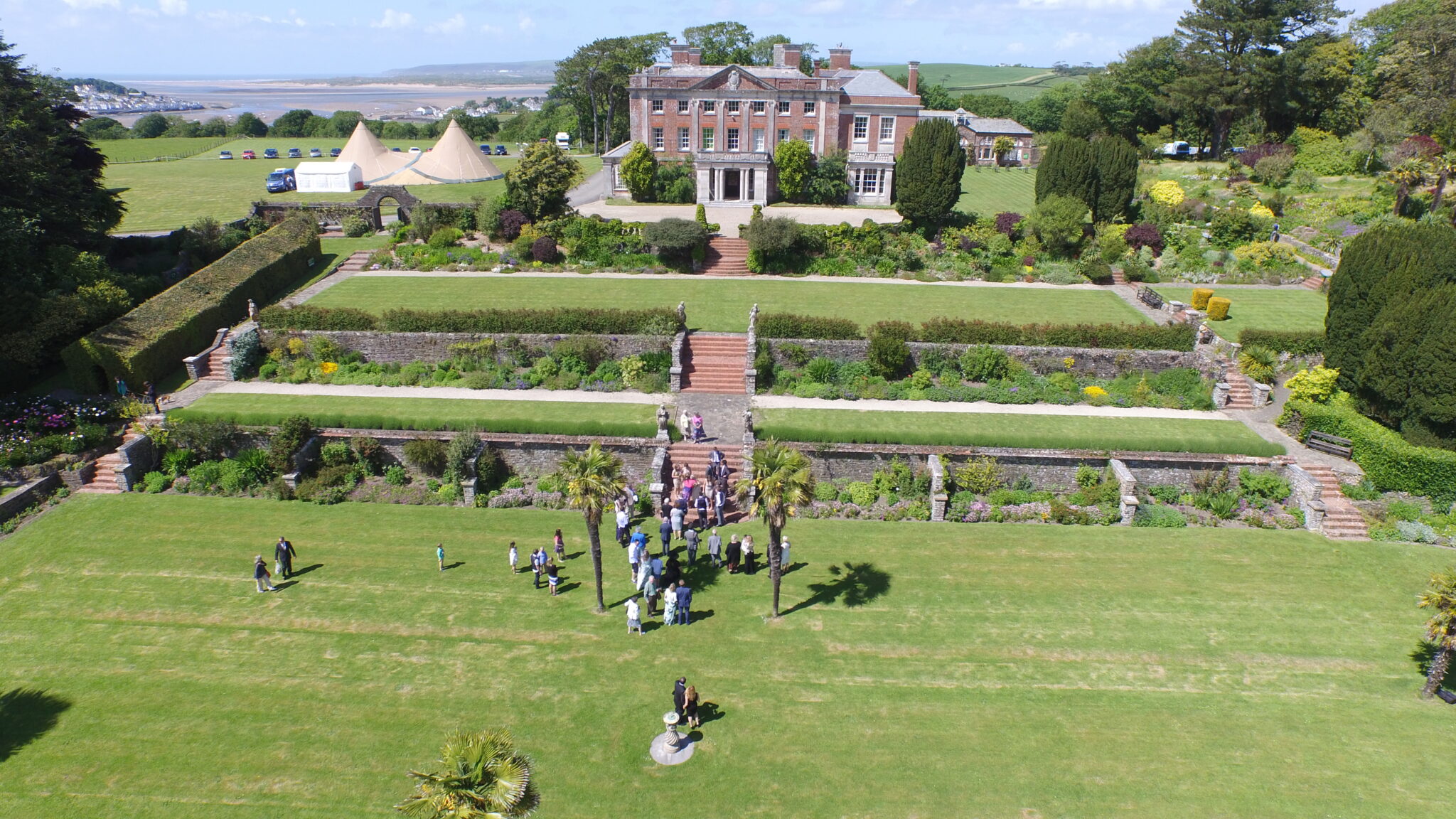 a large stately home type building, with large formal tiered gardens in front of it, leading to a large lawn area