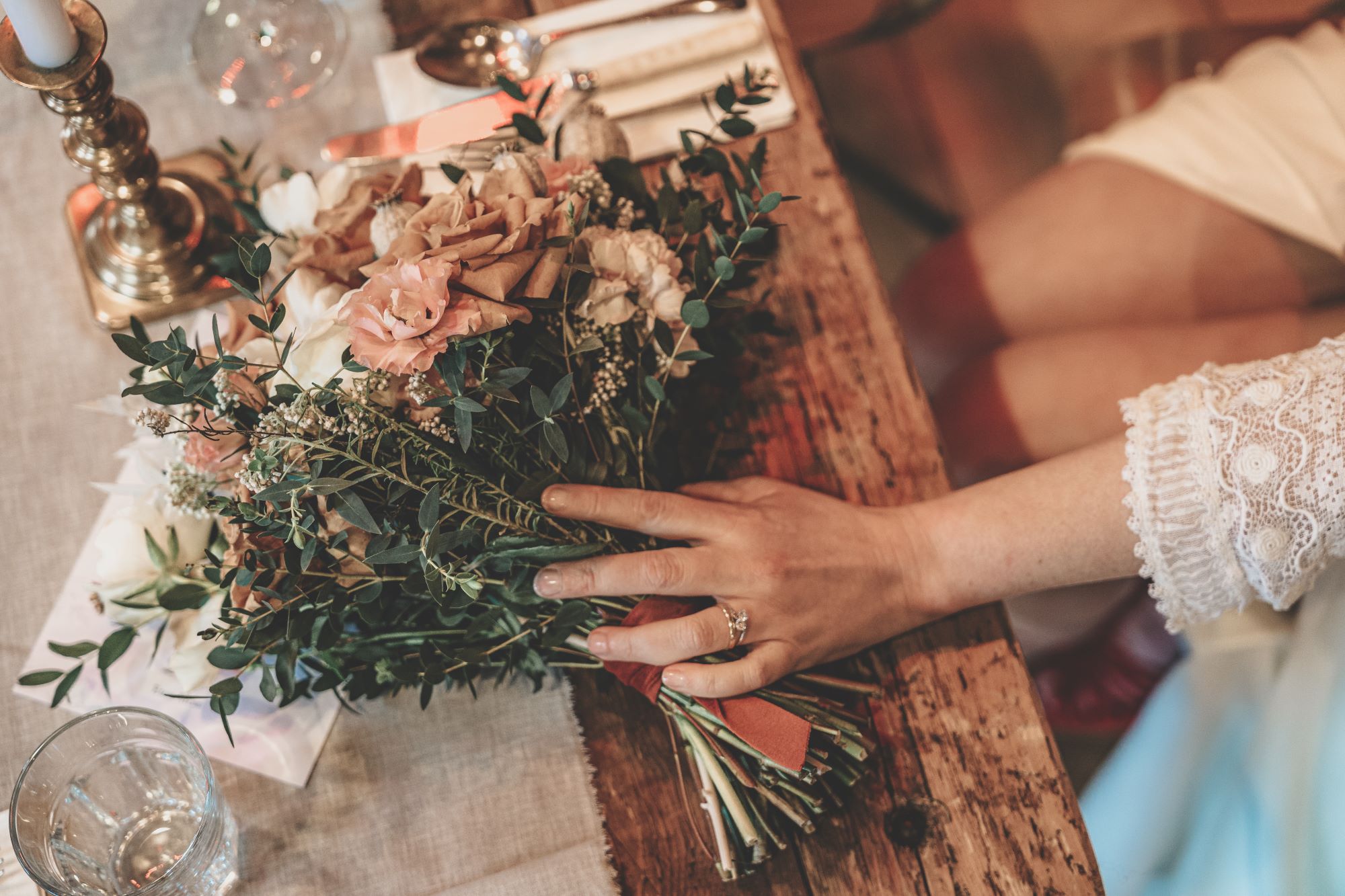 A bride places her hand on a bouquet of flowers that are sat on top of a table at a wedding.