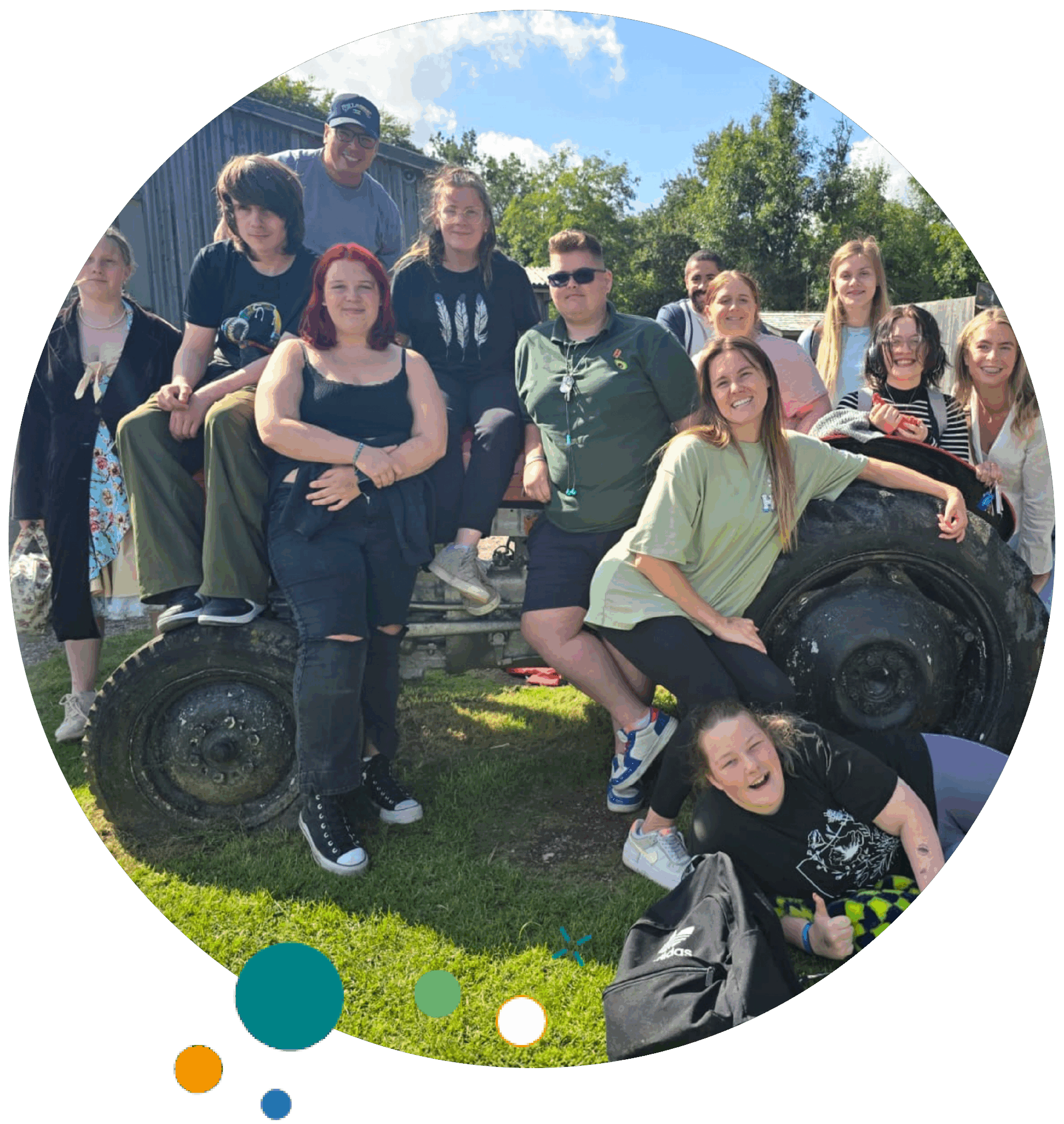 A group of young people and adults posing together outdoors on and around an old tractor wheel. Everyone is smiling, enjoying the sunny day, with trees and a shed in the background.