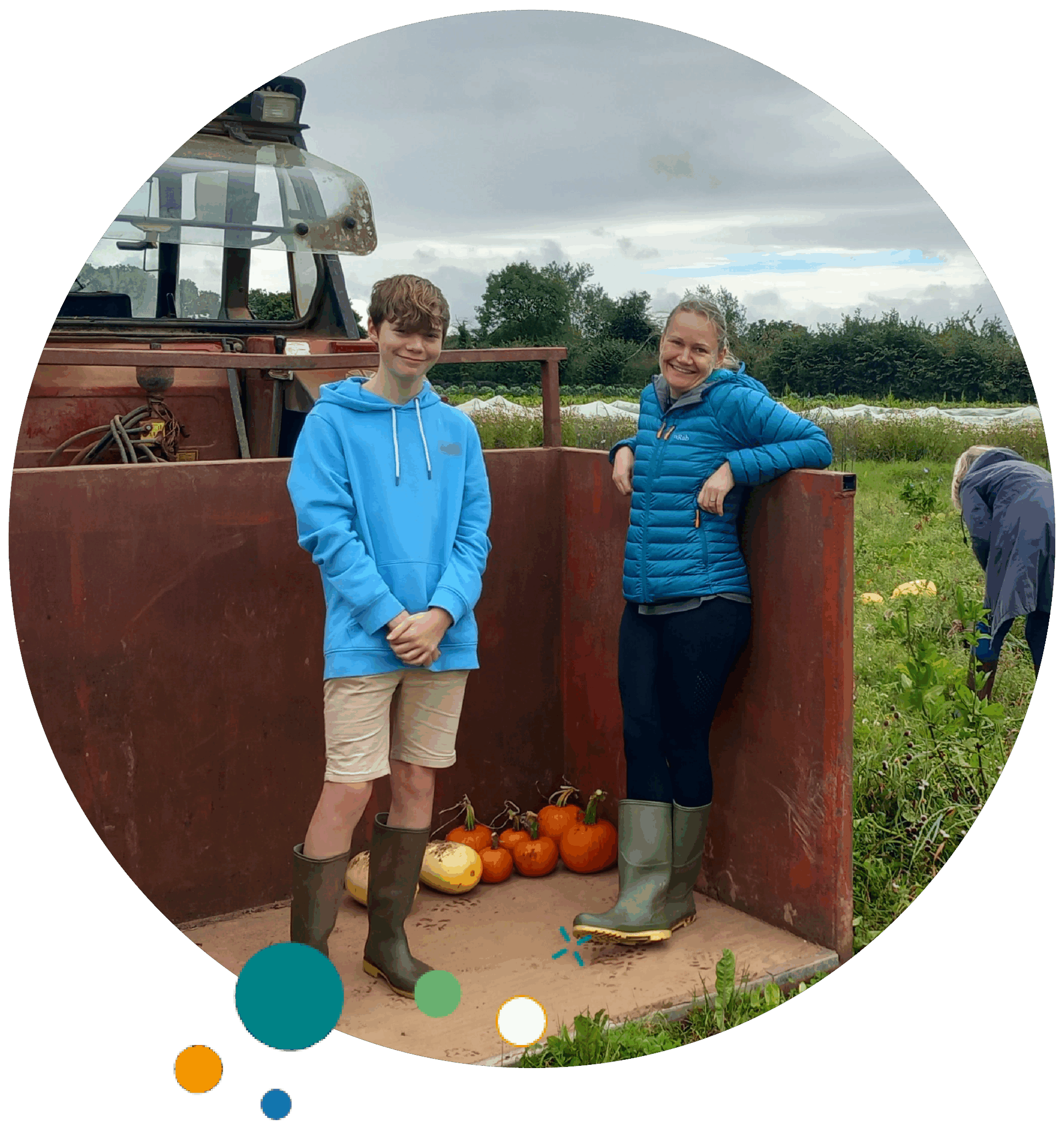 People on a farm during pumpkin harvesting. A boy in a blue hoodie and shorts with green boots stands next to a woman in a teal jacket and black pants inside a tractor trailer with pumpkins on the floor. Other people in the background are picking pumpkins in the field.