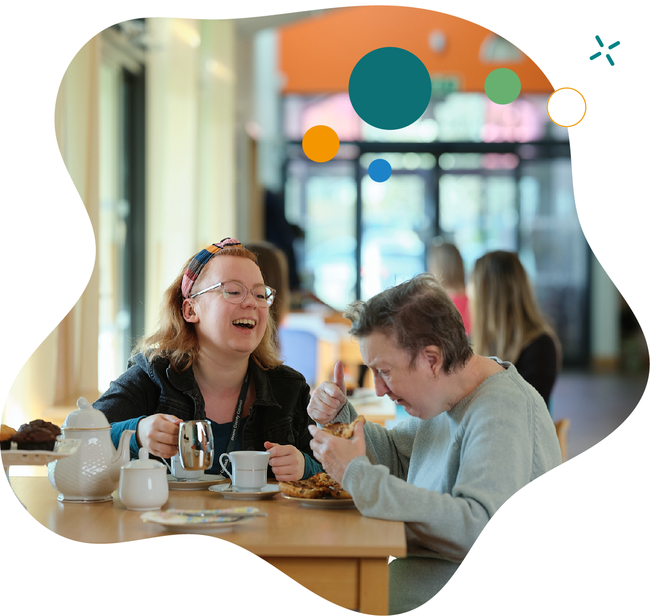 Two women sit together at a table in a café, laughing and enjoying tea and pastries, creating a warm and friendly atmosphere