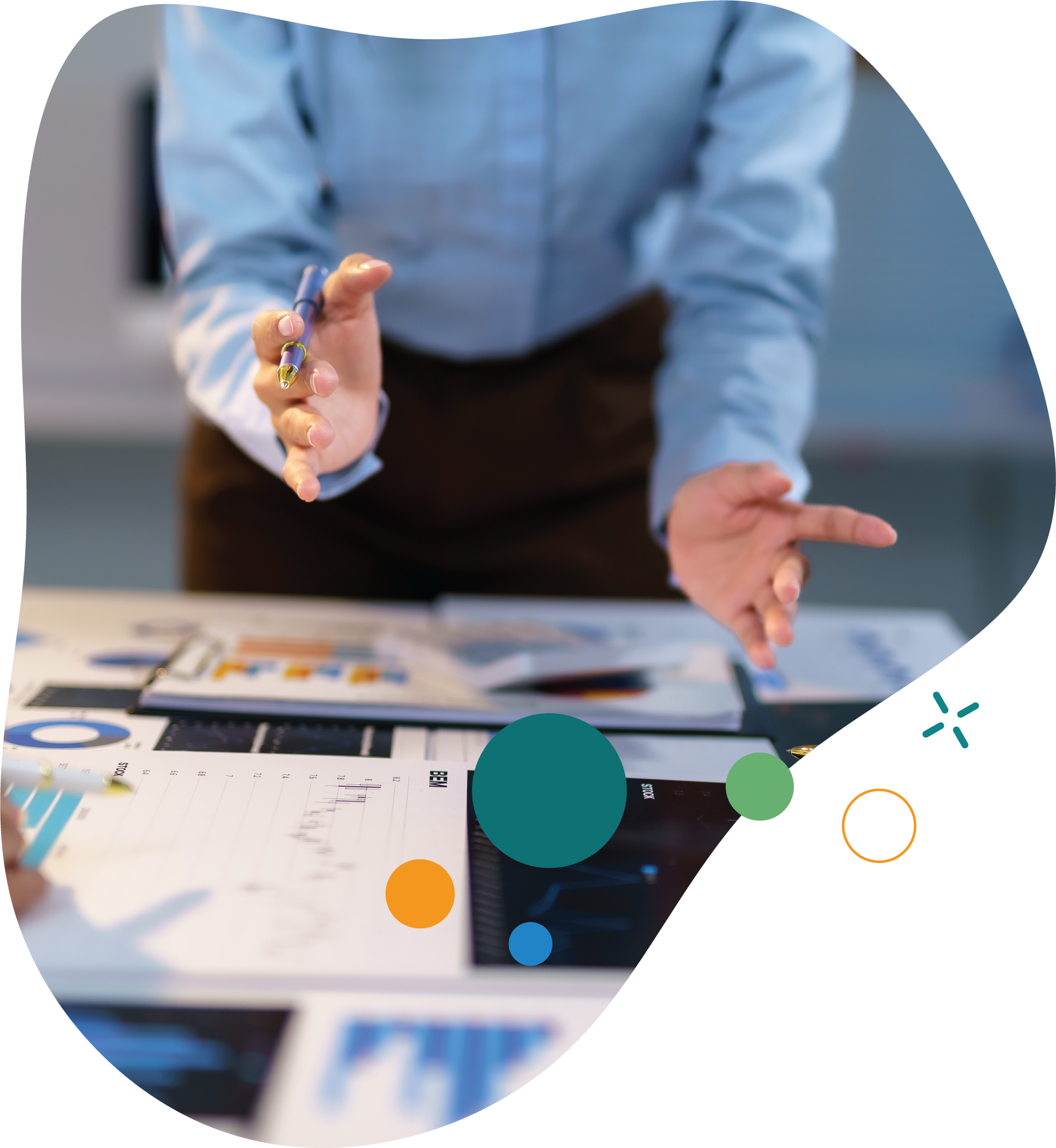 ose-up of a person in a blue shirt gesturing with one hand while holding a pen in the other, leaning over a table covered with charts, graphs, and business documents.