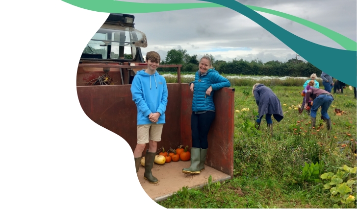 People on a farm during pumpkin harvesting. A boy in a blue hoodie and shorts with green boots stands next to a woman in a teal jacket and black pants inside a tractor trailer with pumpkins on the floor. Other people in the background are picking pumpkins in the field.
